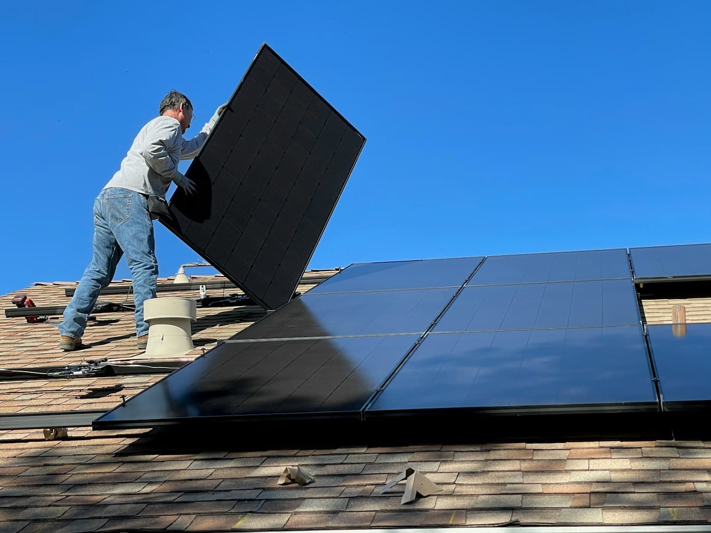 An installer lifting a solar panel onto a shingled roof under a blue sky