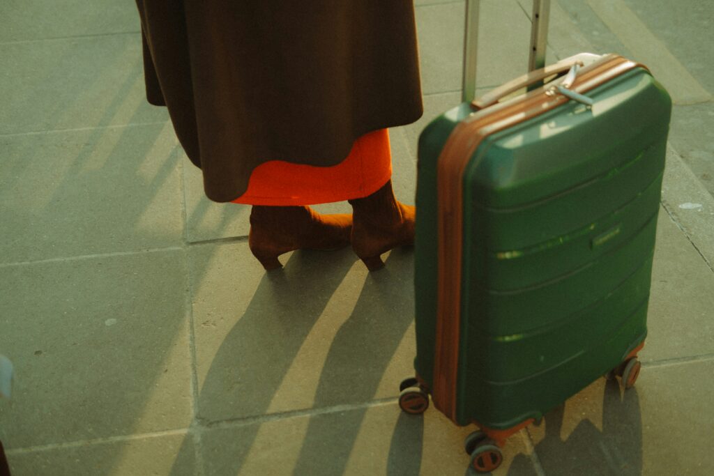 Person in a brown coat and boots stands next to a green rolling suitcase on a sunlit pavement.