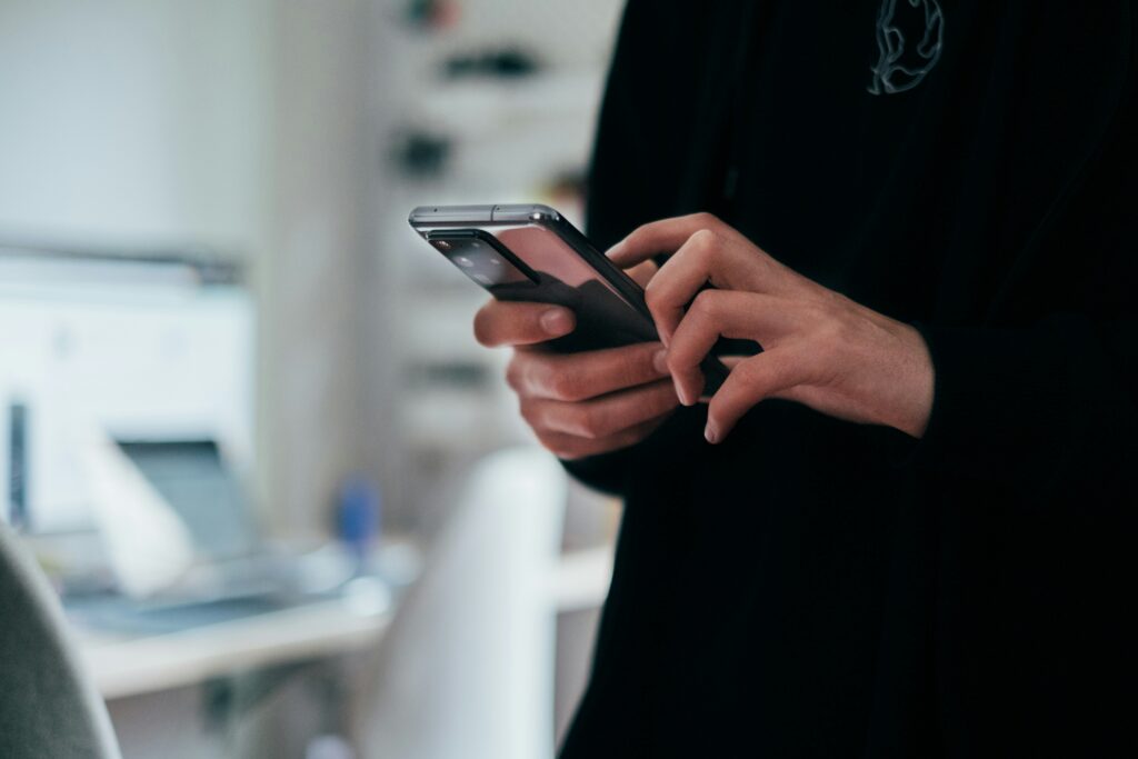 Person holding and using a smartphone indoors, with a blurred background of a desk and computer.