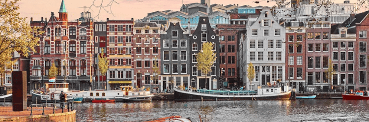 A row of colorful, traditional buildings lines a canal in Amsterdam with boats moored along the water and trees scattered on the banks.