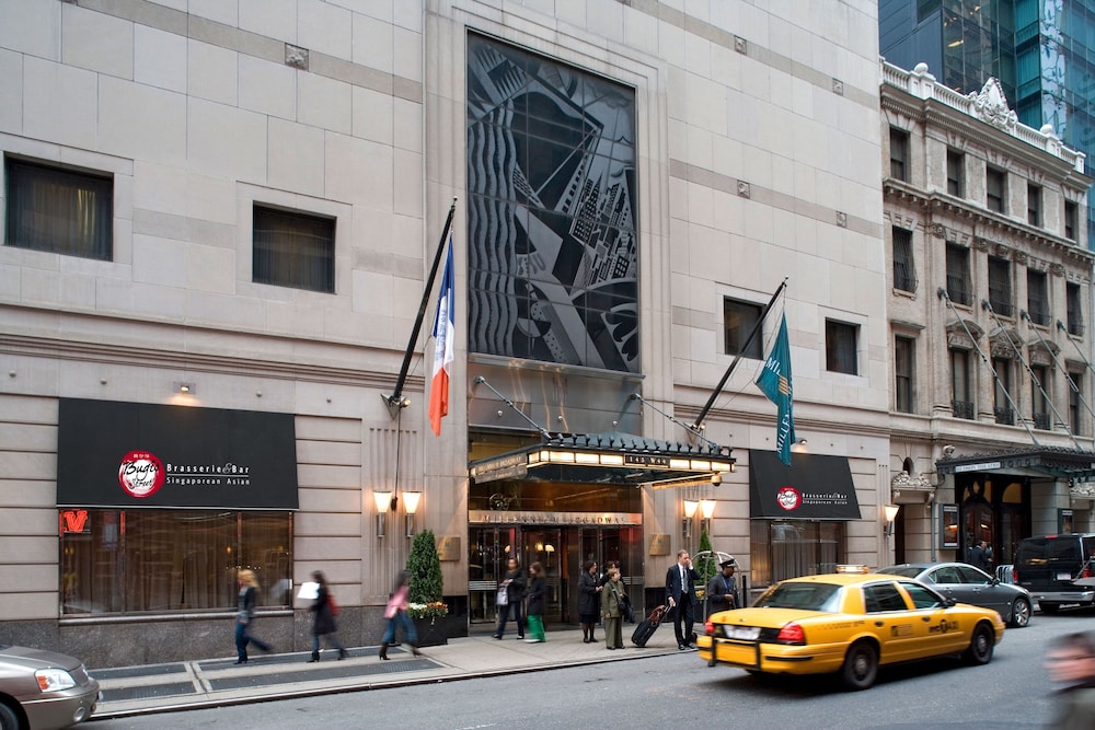 People walk past a hotel entrance with flags above, next to a restaurant and a yellow taxi on the street.