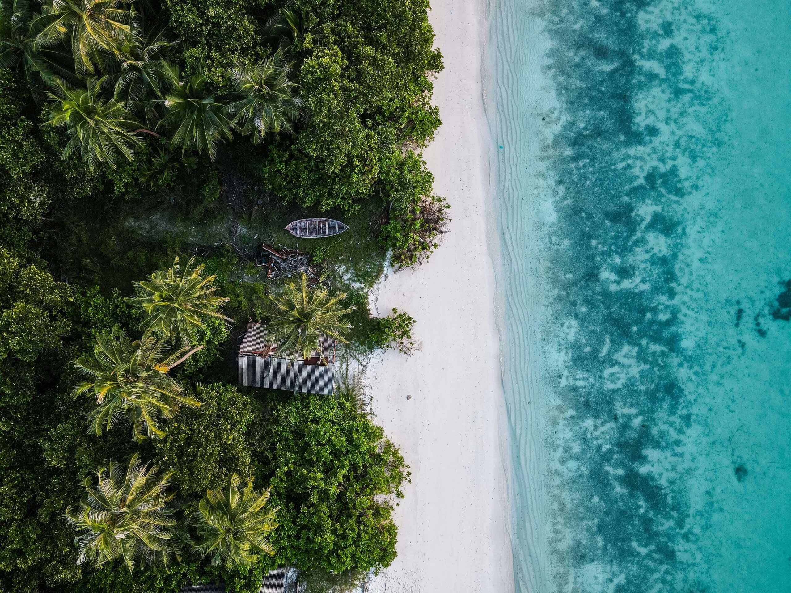 Aerial view of a tropical beach with turquoise water, white sand, lush trees, a small boat, and a hut.