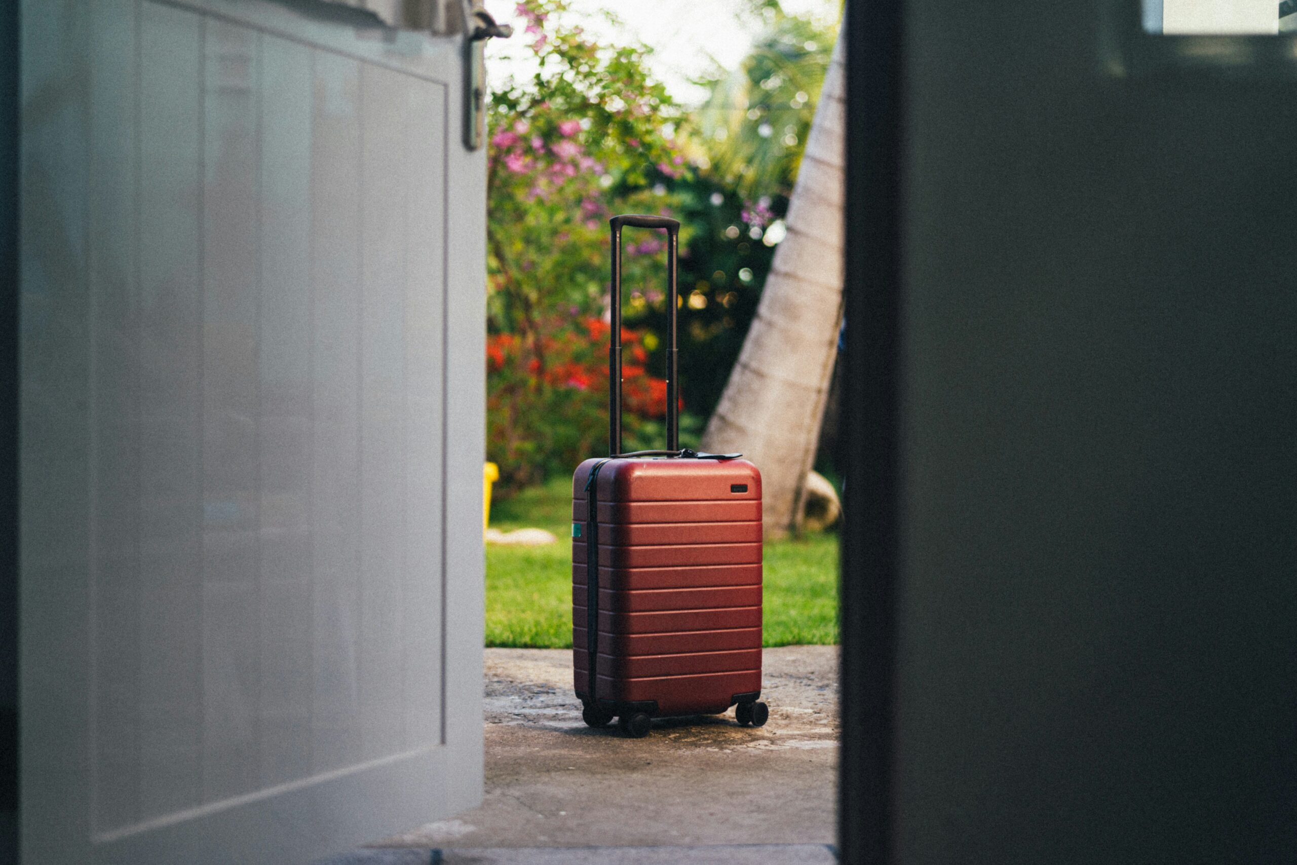 A red suitcase with an extended handle sits outside an open door on a sunny day.