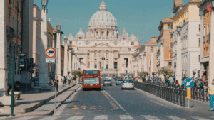 A busy street leading to St. Peter’s Basilica in Vatican City, with cars, buses, and people walking.