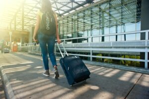Woman walking outside an airport terminal, pulling a suitcase and wearing a backpack in sunlight.