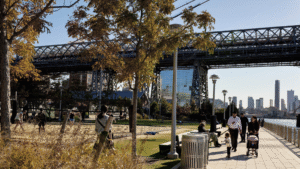 People walk and relax by the river under a large bridge on a sunny day with city buildings in the background.