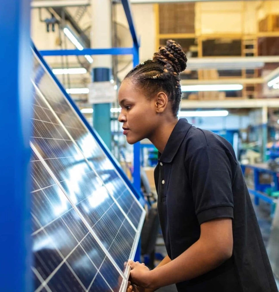 A woman in a black polo shirt inspects a solar panel in an industrial setting.