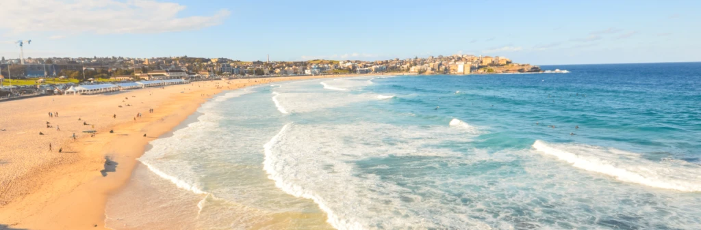 Wide sandy beach with rolling waves, people on the shore, and a coastal town in the distance under a blue sky.