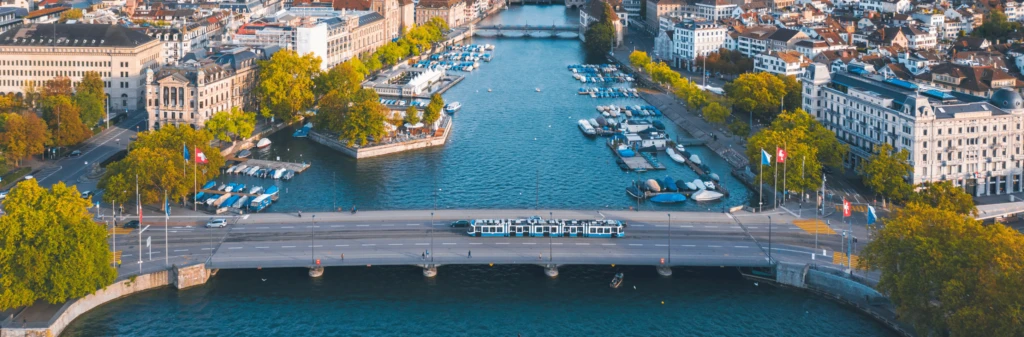 Aerial view of a bridge, river, and boats surrounded by city buildings and trees in Zurich, Switzerland.