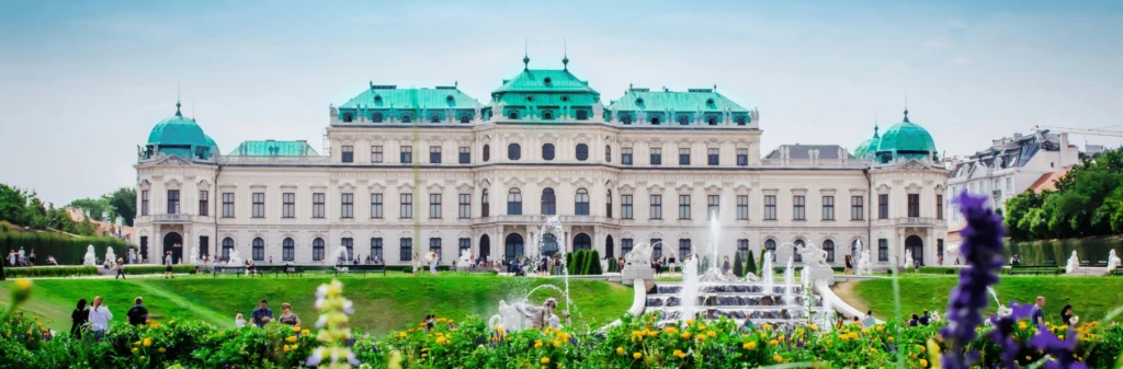 A grand palace with green roofs, gardens, and fountains, crowded with visitors on a sunny day.