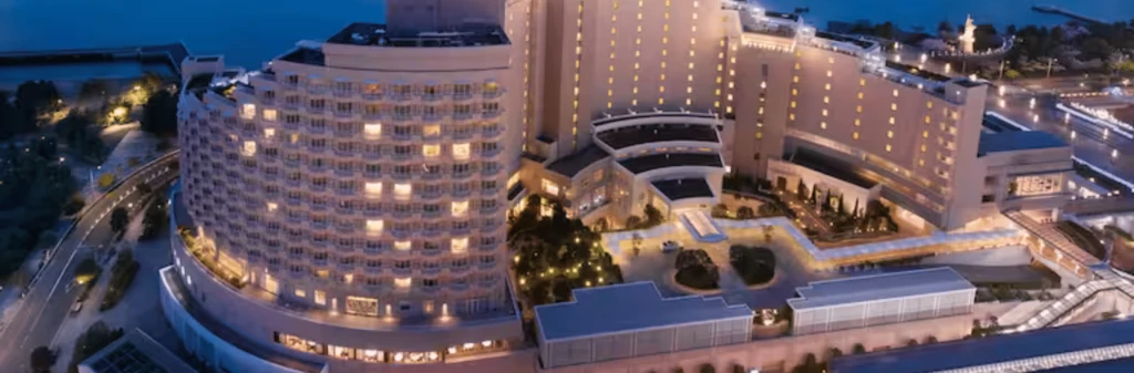 A modern, curved hotel building at dusk with many lit windows and landscaped grounds.
