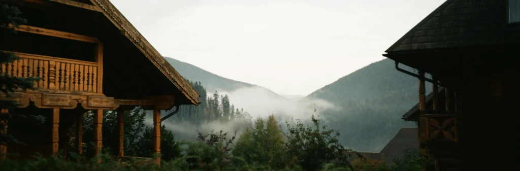 Wooden cabins frame misty forested mountains under a cloudy sky.