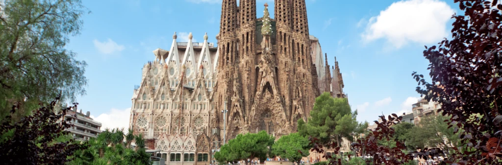 La Sagrada Família basilica in Barcelona, surrounded by trees and people, under a bright blue sky.