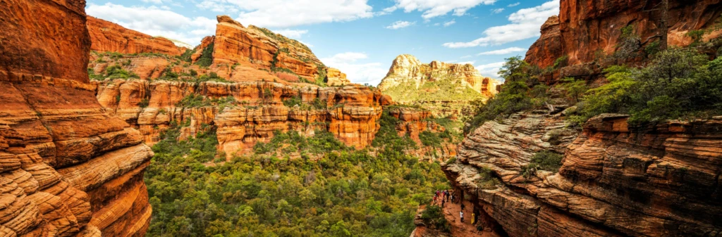 Red rock formations and green trees under a blue sky with clouds in a canyon landscape.