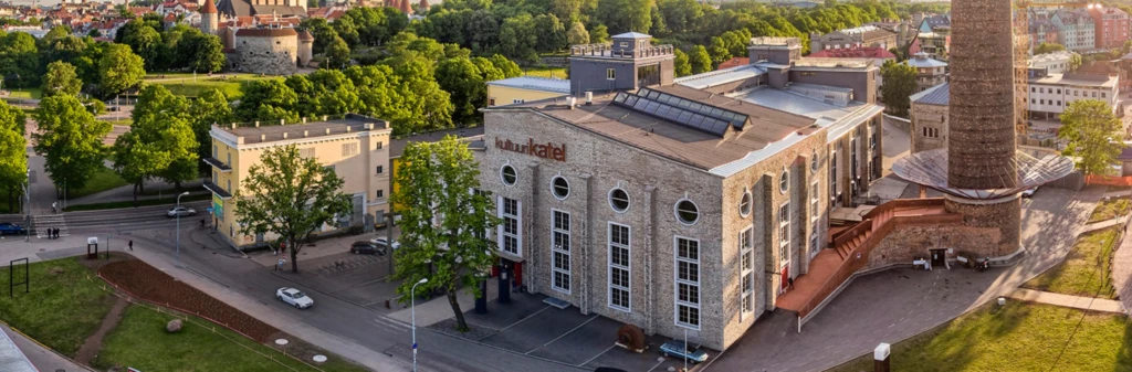 Aerial view of a large brick building with arched windows, tall chimney, and surrounding greenery.