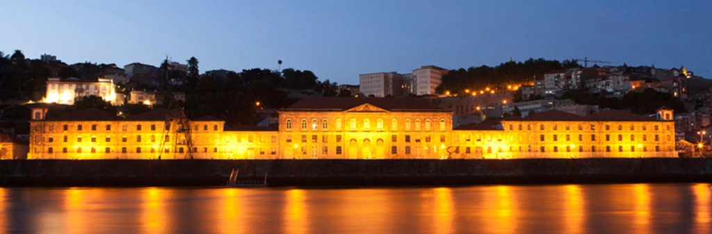 Large illuminated building at night by a river, with hills and city lights in the background.