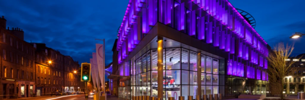 Modern glass building at night, illuminated with vibrant purple lights, on a city street corner.