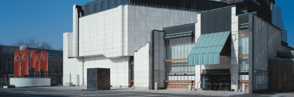 Modern white theater building with angular shapes and glass entrance, red banners on the left, blue sky overhead.