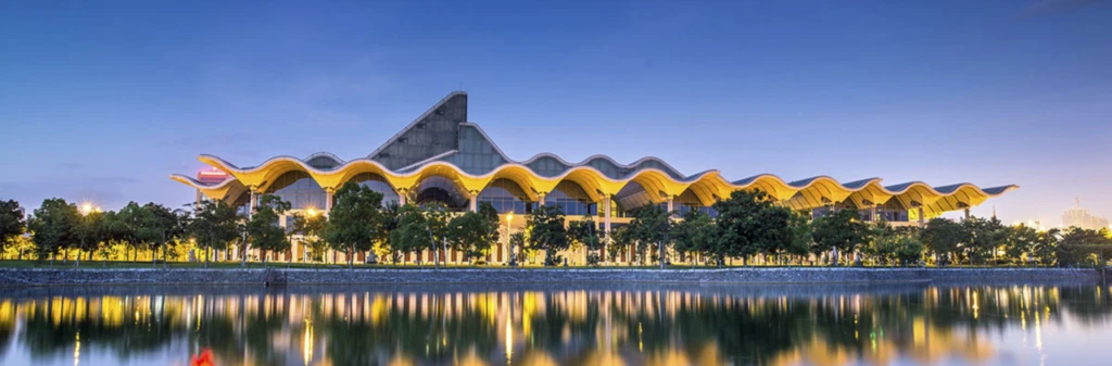 Modern building with a wavy yellow roof, reflected in a calm river, with trees and lights at dusk.