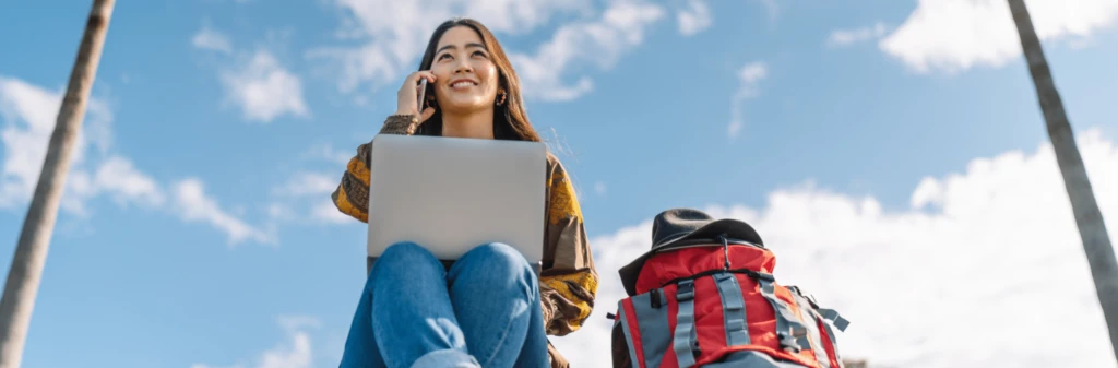 Smiling woman outdoors with laptop, talking on phone, sitting next to a backpack under a blue sky with clouds.