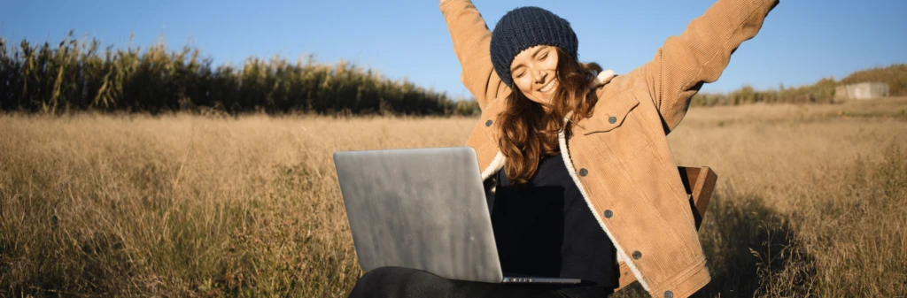 Woman sitting outdoors with a laptop, smiling and stretching arms up in a field under a clear blue sky.