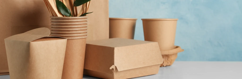 Assorted brown eco-friendly paper food containers, cups, and utensils on a white surface with a blue background.