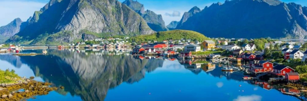 Colorful houses by a calm lake, with tall mountains reflected in the water under a clear blue sky.