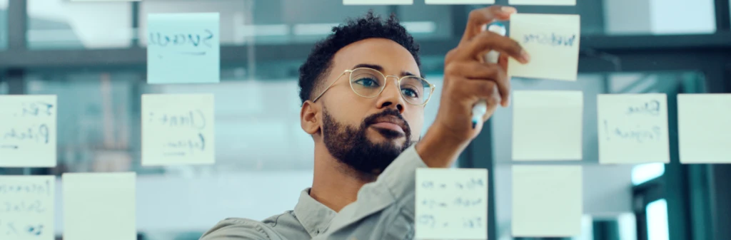 Man writing on sticky notes on a glass wall, surrounded by other notes in an office setting.