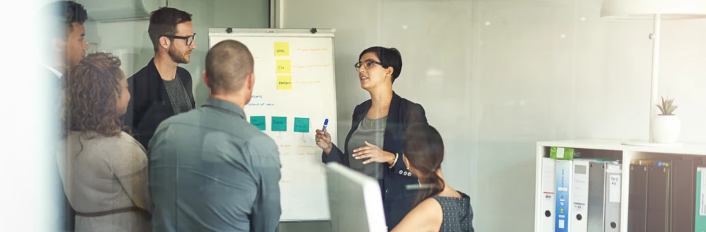 A woman leads a meeting, presenting ideas on a flipchart to a group in a modern office.