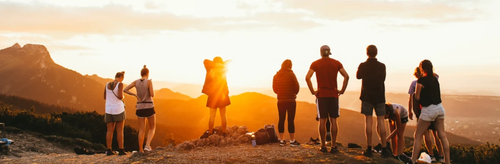 A group of people stand on a mountaintop at sunset, overlooking scenic mountains and valleys.