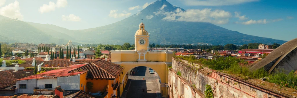 Yellow clock arch in Antigua, Guatemala, with a volcano and blue sky in the background.