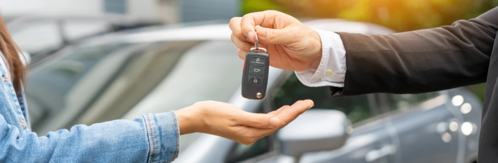 A person hands car keys to another person standing beside a parked car outdoors.