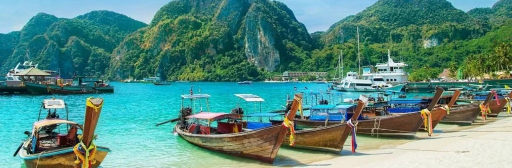 Long-tail boats and yachts float on clear turquoise water with green mountains in the background.