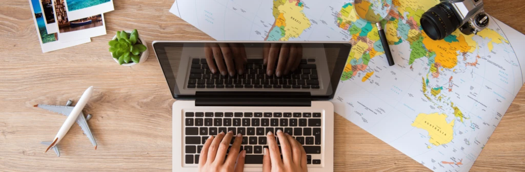 Hands typing on a laptop next to a map, camera, miniature airplane, plant, and travel photos on a desk.