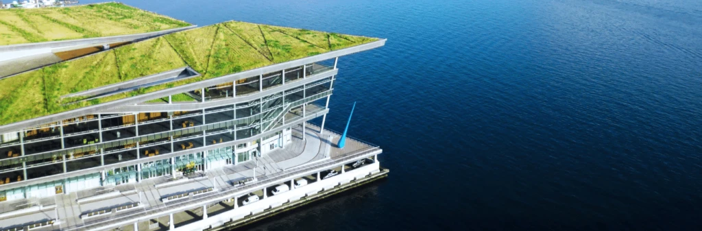 Modern glass building with a green rooftop next to calm blue water, viewed from above.