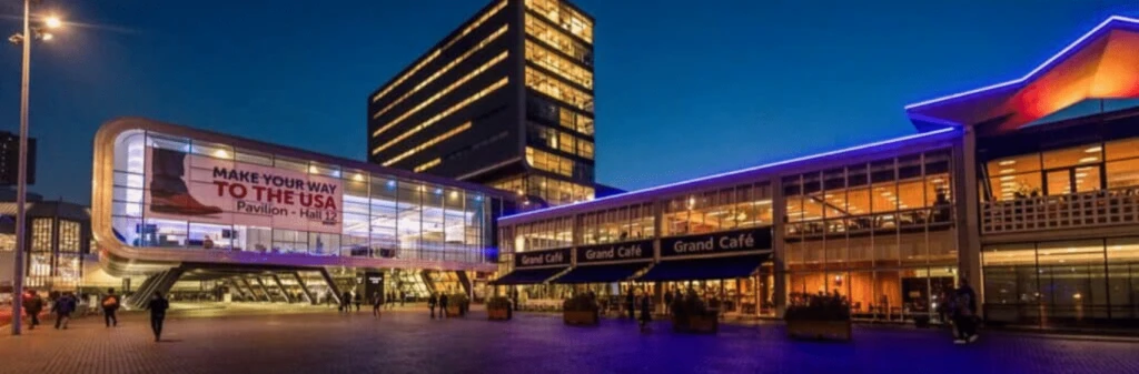 Modern buildings lit up at night with a large Make Your Way to the USA banner and people walking outside.