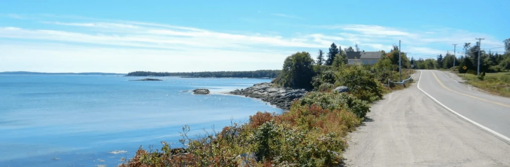A coastal road curves beside calm blue water, with trees and a house in the distance under a clear sky.