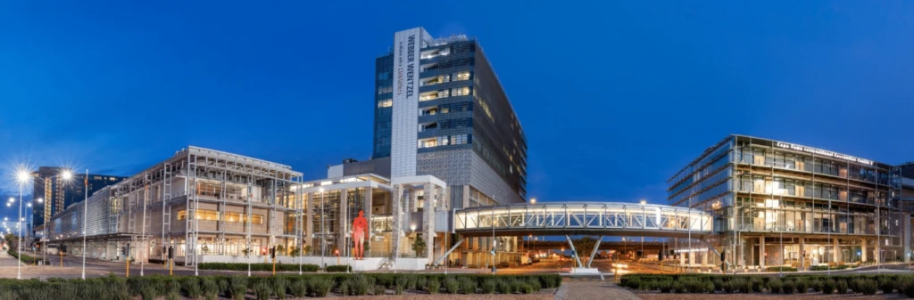 Modern university buildings and pedestrian bridge lit up at dusk, with clear blue evening sky in the background.