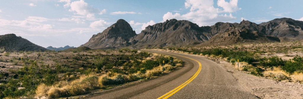 A winding road curves through a desert landscape with rocky mountains under a partly cloudy sky.