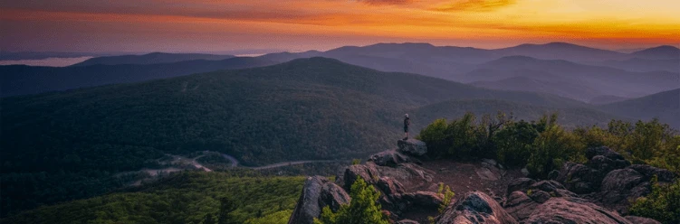 Person stands on rocky overlook, gazing at rolling green hills under a colorful sunset sky.