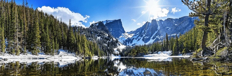 Mountain lake with snowy peaks, pine trees, and bright sunlight reflecting on the water under a blue sky.
