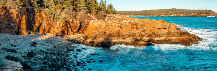 Rocky coastline with orange cliffs, scattered trees, and waves crashing onto the shore under clear skies.
