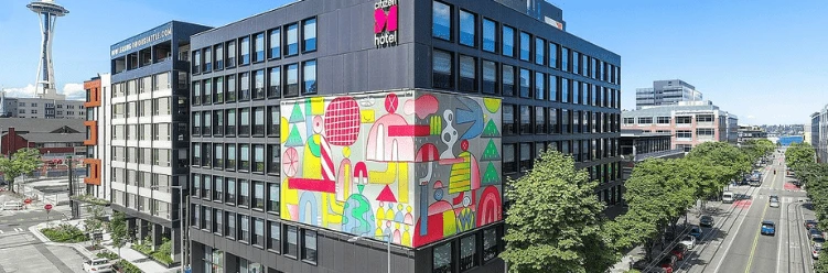 A modern multi-story hotel with a colorful geometric mural on the corner, located on a city street with the Space Needle visible.