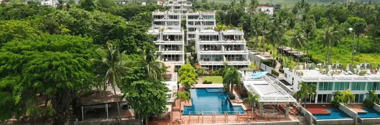Aerial view of a modern multi-story resort complex with swimming pools, surrounded by lush greenery and palm trees.