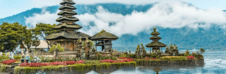 Balinese temple by a lake with mountains and clouds in the background, surrounded by flowers and greenery.