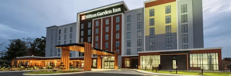 Exterior view of a Hilton Garden Inn hotel, featuring modern architecture with large windows and illuminated signage at dusk.
