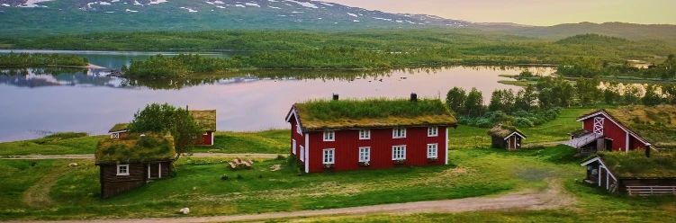 Red houses with grass roofs by a lake and mountains, surrounded by lush green landscape at sunset.