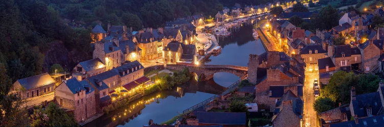 A scenic riverside village at dusk, with stone houses, a stone bridge, and warm glowing streetlights.