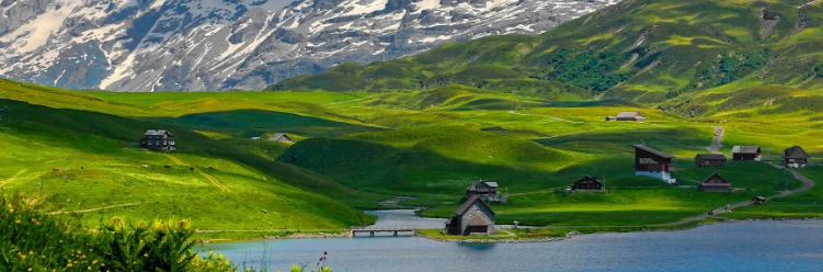 Traditional Swiss chalets near a calm lake, surrounded by rolling green hills and snow-covered Alpine peaks in the background.
