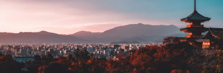 Panoramic view of Kyoto at sunset, with a traditional Japanese temple in the foreground, autumn trees, and mountain silhouettes in the distance under a colorful sky.
